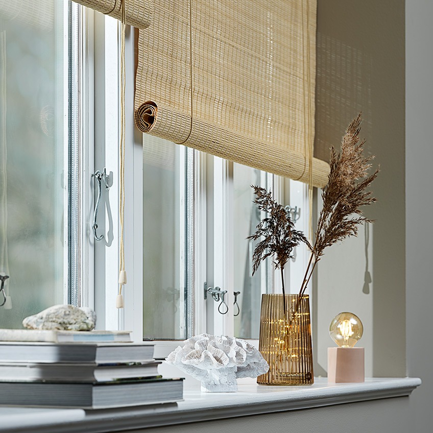 Rolety bambusowe w oknie z doniczką, ornamentem, wazonem, świecą zapachową i lampką bateryjną w parapecie