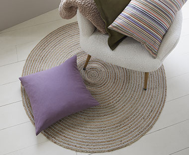 Okrągły dywan, na którym stoi jasny fotel i leży fioletowa poduszka