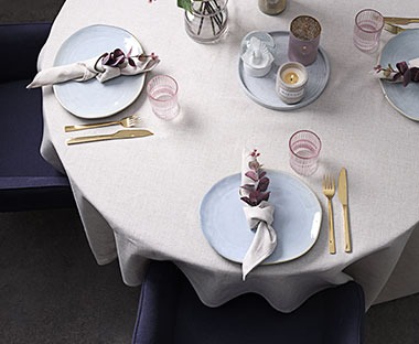 Okrągły stół z jasnoszarym obrusem oraz zastawą z niebieskich talerzy i złotych sztućcy