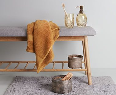 Pomarańczowy ręcznik, który jest przewieszony na szarej ławce, na której stoją akcesoria łazienkowe