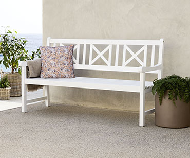 Biała ławka ogrodowa ustawiona pod ścianą na której leży różowa poduszka 