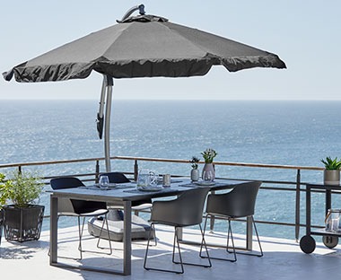 Parasol ogrodowy otwarty na tarasie z widokiem na morzem pod którym stoi zestaw mebli ogrodowych złożony ze stoły i 4 krzeseł 
