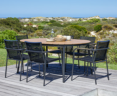 Owalny stół ogrodowy z czarnymi nogami i drewnianym blaem oraz czarne krzesła na tarasie z widokiem na zieleń i morze 