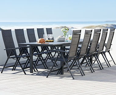 Duży, czarny stół na podeście nad brzegiem morza z 8 krzesłam z wysokimi oparciami. 