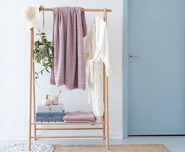 Jasny, drewniany stojak na ubrania na tle białej ściany obok błękitnych drzwi. 