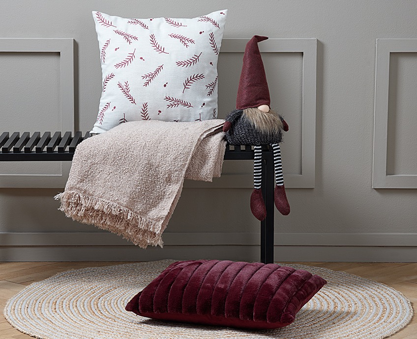 Ławka z poduszką, kocem i elfem. Na podłodze przed ławką czerwona poduszka na okrągłym dywanie