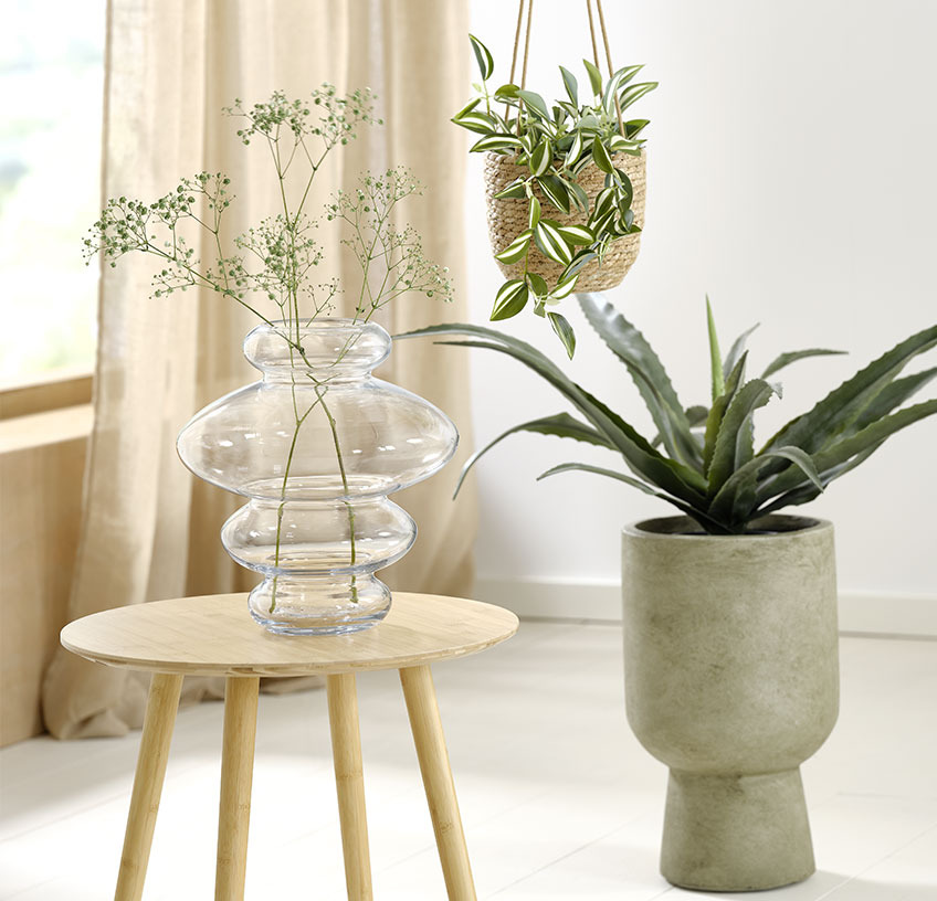 : szklany wazon na stoliku, wisząca doniczka i zielona, duża donica ze sztucznym kwiatem. 