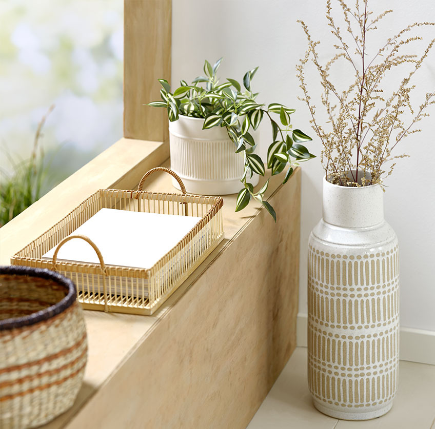 Wysoki wazon obok parapetu, na którym leży bambusowa taca i biała doniczka ze sztucznym kwiatem 