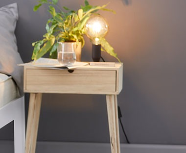 Drewniany stolik nocny z szufladą i kwiatkiem oraz lampką położonymi na nim. 