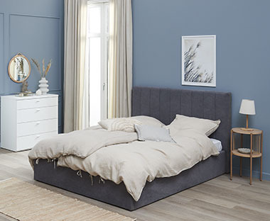 Szare, tapicerowane łóżko w niebieskiej sypialni z beżową pościelą 