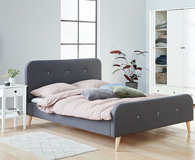 Szare łóżko na drewnianych nóżkach w jasnej sypialni 