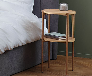 Drewniany stolik z okrągłym blatem i półką obok szarego łóżka z białą pościelą 